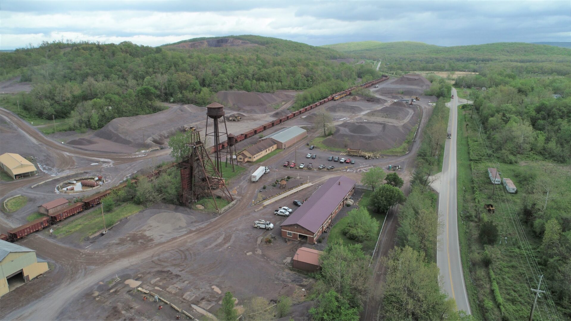 Aerial view of the Iron Mountain property, April 2020: the rusted steel headframe and water tower at center, the long red-roofed building in the foreground, ore cars on the internal rail line, stockpiles of crushed trap rock, a circular basin at far left, and the wooded mountain rising behind.