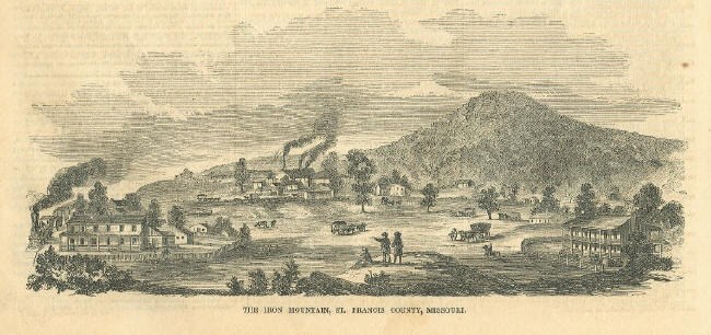 An 1857 engraving of Iron Mountain, St. Francis County, Missouri: the wooded mountain rising behind a company town of frame buildings, with smokestacks trailing smoke and figures and wagons on the road in the foreground.