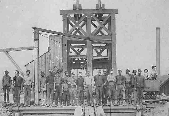 A mine crew of roughly twenty men — and one boy — posed in a line before a timber headframe, with an ore car on rails at right and a smokestack behind.
