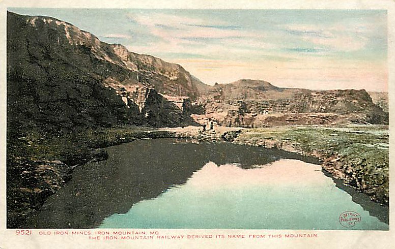 A hand-tinted postcard of the old iron mines at Iron Mountain: water-filled workings reflecting the sky beneath weathered rock walls, captioned that the Iron Mountain Railway derived its name from this mountain.