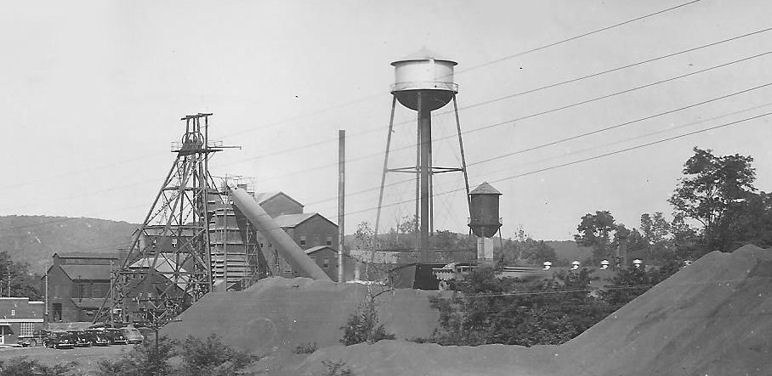 The Ozark Ore Company surface plant at Iron Mountain, Missouri: the headframe at left beside the mill buildings, a tall water tower at center, and ore piles in the foreground, photographed in the mid-twentieth century.